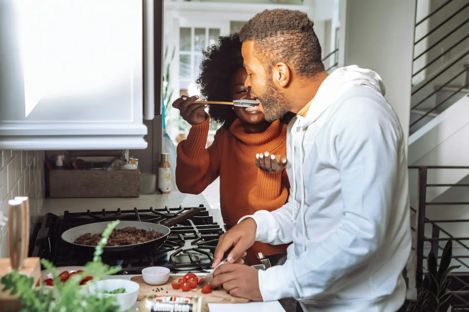 A couple cooking together, one giving the other a taste from a spoon