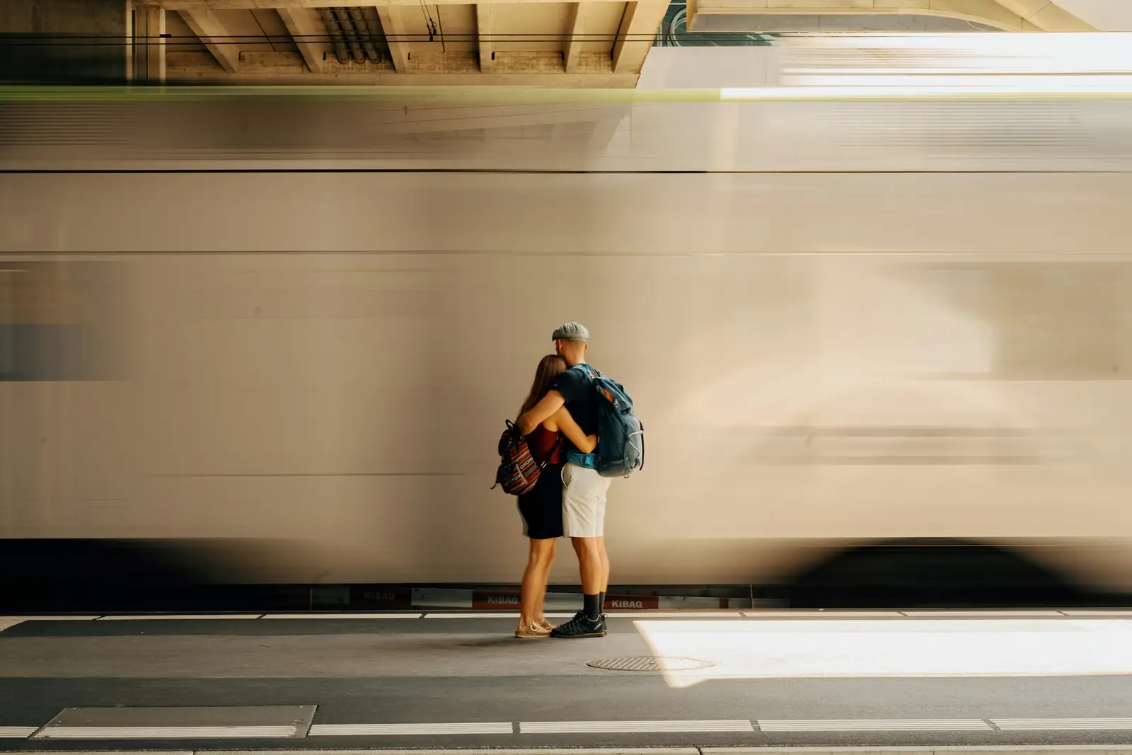 Two people with backpacks hugging on a station platform as a train rushes past